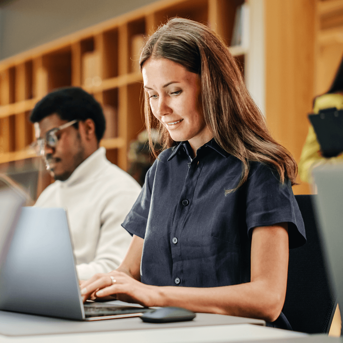 Woman working on a laptop at a desk in a modern office, with a colleague in the background.