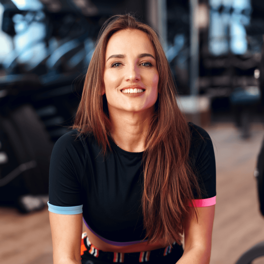 Smiling woman in athletic wear seated in a gym setting, with workout equipment blurred in the background.