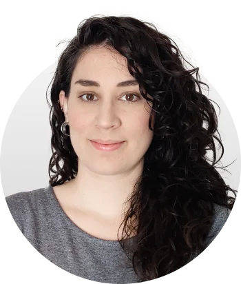 Woman with long dark curly hair looks directly at camera with a gentle smile, wearing a gray top and small hoop earrings against a light gray...