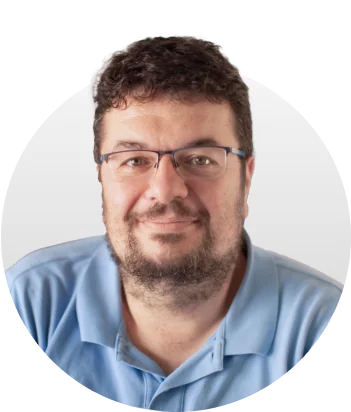 Adult man with glasses and short dark hair smiles slightly while facing the camera; light stubble and beard, wearing a light blue polo against a pale...