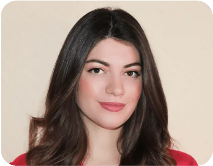 Young woman gazes at the camera with a gentle smile, long dark wavy hair and natural makeup, wearing a red top against a softly lit beige background.