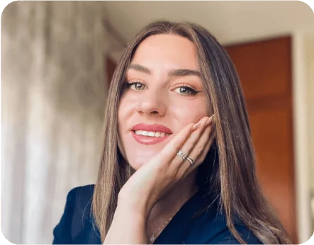 Young woman smiles, resting her chin on her hand; indoors with blurred curtains and a wooden door behind her, wearing a dark top and ringed fingers.