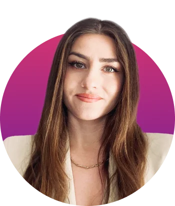 Young woman looks into camera, smiling slightly, wearing a beige blazer and thin gold chain necklace; long brown hair frames her face against a...