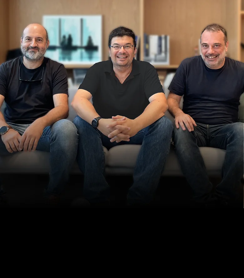 Three men sit on a couch, smiling; center man clasps his hands, others rest on knees, in a wood-paneled lounge with blurred artwork and shelves...