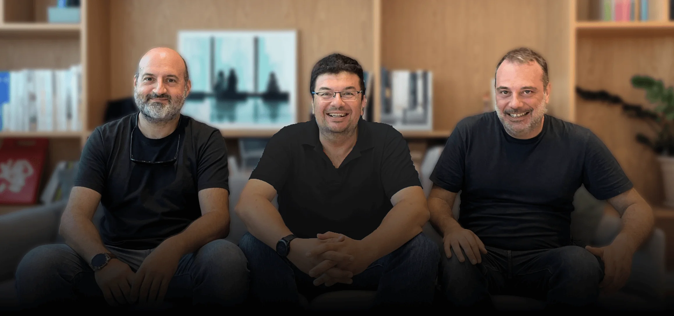 Three men sit smiling at the camera on a sofa, hands clasped; they wear dark shirts; background shows wooden bookshelves, books, and framed artwork.