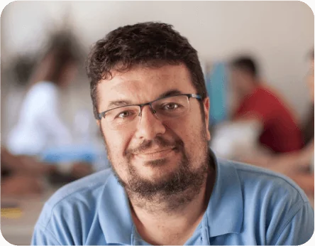 Bearded man wearing glasses and a blue polo smiles at the camera, seated in a softly lit indoor space with blurred people and tables in the...