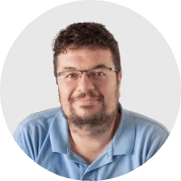 Bearded man with glasses wearing a light-blue polo smiles gently at the camera, head-and-shoulders portrait framed within a circular light-gray...