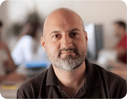 Bald, bearded man looks directly at camera with a neutral expression, seated in a softly lit open office with blurred coworkers and computer screens...